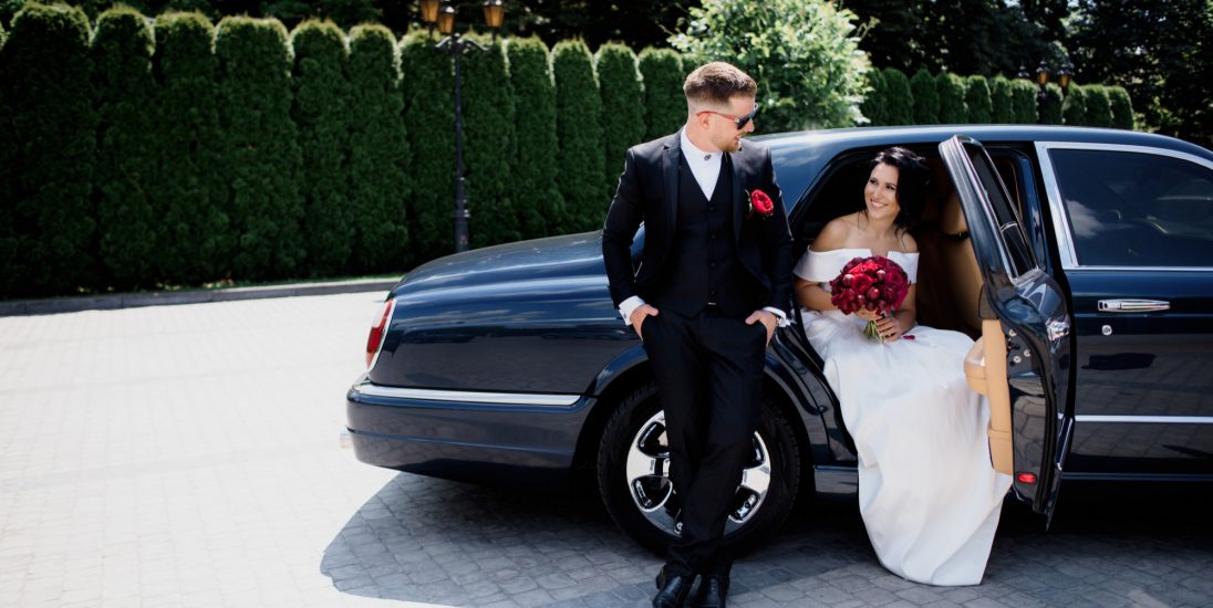 beautiful-wedding-couple-is-smiling-in-the-black-car-on-the-sunny-day-dressed-in-elegant-wedding-outfits-with-red-bouquet