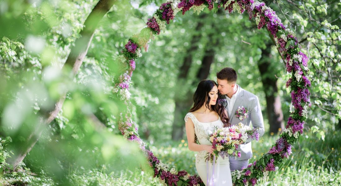 bride-and-groom-pose-behind-large-circle-of-lilac-in-the-garden